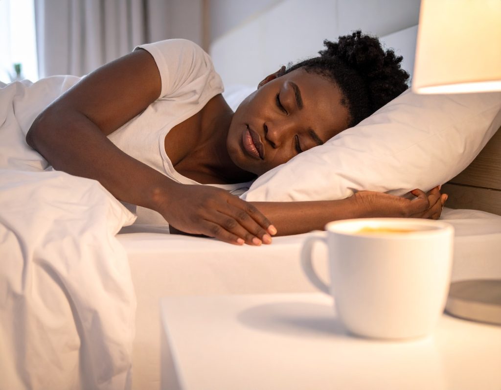 A steaming cup of coffee next to a person peacefully napping in bed