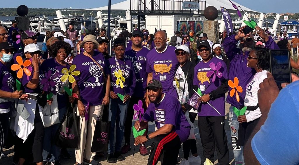 Congressman Glenn Ivey walks with families at the Alzheimer’s Walk at National Harbor, surrounded by purple flowers and tribute signs.